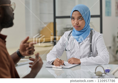 Young female doctor in hijab and uniform attentively listening to her patient and taking notes 110957786