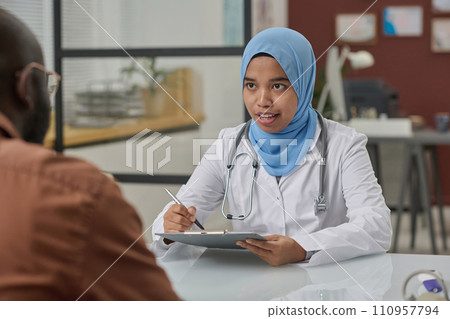 Female doctor in hijab talking to her black patient and writing information on paper on clipboard 110957794