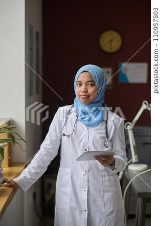 Female physician in hijab and lab coat confidently posing at hospital office holding digital tablet 110957803