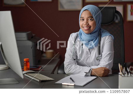 Portrait of female medical worker in hijab and uniform sitting at desk in clinic smiling at camera 110957807