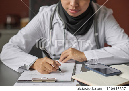 Unrecognizable young female doctor filling documents sitting at desk in hospital office 110957837
