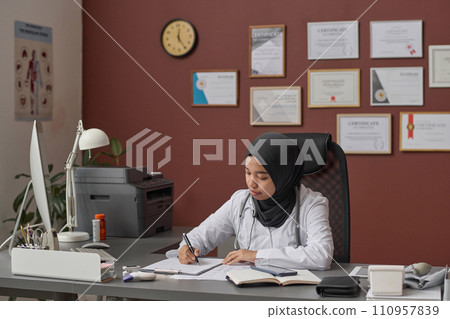 Young female doctor in hijab sitting at clinic desk and attentively filling documents 110957839