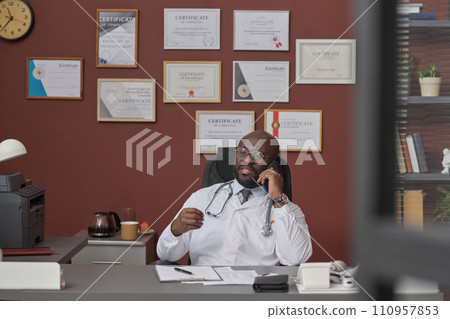 African american doctor talking on his smartphone while sitting at his workplace in clinic 110957853