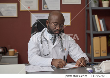 African american doctor attentively writing on paper attached to clipboard sitting at his workplace African american doctor attentively writing on paper attached to clipboard sitting at his workplace 110957869