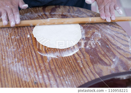 Close up of old female coock, chef wearing apron, rolling dough on wooden board dusted with flour. Concept of fast food. Close up of old female coock, chef wearing apron, rolling dough on wooden board dusted with flour. Concept of fast food. 110958221