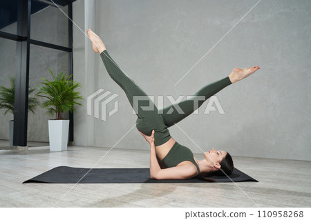 Young slender woman in tight leggings practicing yoga legs up pose on yoga mat at home. Side view of focused girl doing hatha yoga postures in modern indoor space. Concept of yoga practice, sport. Young slender woman in tight leggings practicing yoga legs up pose on yoga mat at home. Side view of focused girl doing hatha yoga postures in modern indoor space. Concept of yoga practice, sport. 110958268