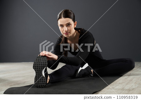 Beautiful focused woman stretching body, while leaning to leg before exercise on yoga mat. Front view of sportswoman wearing sneakers, looking at camera, while stretching ankle. Concept of exercising. 110958319