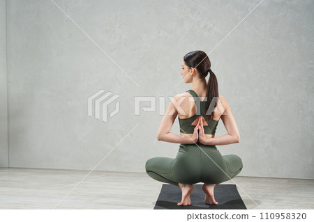 Unrecognizable woman wearing green gym clothes sitting back in yoga asana, with hands in namaste gesture behind back on gray background with studio light. Mindfulness and mental health concept. 110958320