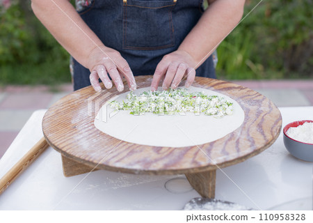 Close up of old female cook, chef wearing apron making pita, putting cheese filling on dough. Concept of fast food. 110958328