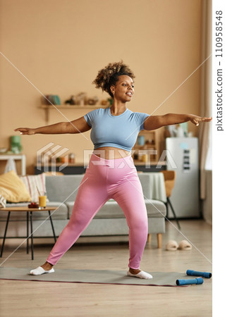 Vertical shot of young African American expectant mother wearing fitness clothes exercising at home while standing in yoga warrior pose with outstretched arms Vertical shot of young African American expectant mother wearing fitness clothes exercising at home while standing in yoga warrior pose with outstretched arms 110958548