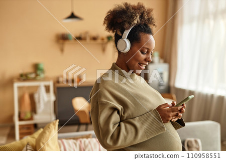 Medium shot of smiling pregnant Black woman at home wearing headphones holding smartphone and texting with smartphone 110958551