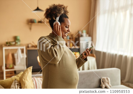 Waist up shot of smiling pregnant Black woman vibing to music looking at smartphone in hand while standing in cozy living room Waist up shot of smiling pregnant Black woman vibing to music looking at smartphone in hand while standing in cozy living room 110958553