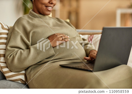 Cropped shot of smiling pregnant Black woman working on laptop while putting hand on her big belly sitting on sofa at home 110958559