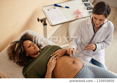 Top down view of pregnant Black woman lying on examination table in medical center exposing big belly while senior doctor listening to baby heart sounds Top down view of pregnant Black woman lying on examination table in medical center exposing big belly while senior doctor listening to baby heart sounds 110958608