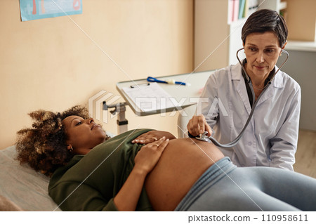 High angle view of senior Caucasian obstetrician using stethoscope listening to baby heartbeat while pregnant Black woman lying on examination bed in hospital High angle view of senior Caucasian obstetrician using stethoscope listening to baby heartbeat while pregnant Black woman lying on examination bed in hospital 110958611