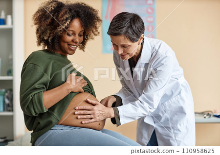 Medium full shot of senior Caucasian female gynecologist using hands for examining cheerful pregnant African American womans uncovered abdomen in clinic Medium full shot of senior Caucasian female gynecologist using hands for examining cheerful pregnant African American womans uncovered abdomen in clinic 110958625