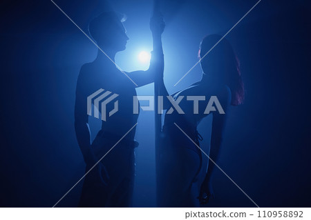 Silhouettes of woman and man dancing on stage holding on to pole illuminated with blue spotlight beam Silhouettes of woman and man dancing on stage holding on to pole illuminated with blue spotlight beam 110958892