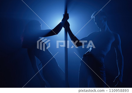 Medium long shot of muscular male pole dancer and female partner performing reverse body waves on stage in blue backlight Medium long shot of muscular male pole dancer and female partner performing reverse body waves on stage in blue backlight 110958893