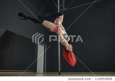 Full shot of strong Caucasian female pole dancer wearing high heel boots doing outside leg hang stunt in studio 110958906