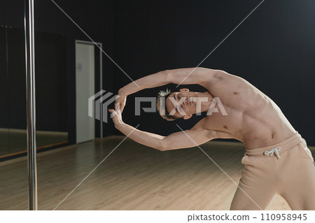 Medium long shot of young Caucasian male pole dancer doing stretching exercise standing in studio 110958945