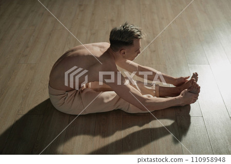 Top down with side view of topless male dancer bending and doing leg muscle stretching exercise sitting on floor Top down with side view of topless male dancer bending and doing leg muscle stretching exercise sitting on floor 110958948