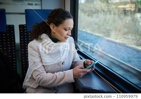 Attractive young adult woman using smart phone while enjoying her business travel by train, sitting on a seat by window overlooking downtown. People. Lifestyle. Public railroad transportation concept 110959979