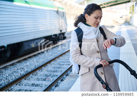 Young mother checks time on wristwatch, standing on railway station platform against a moving train on the background 110959985