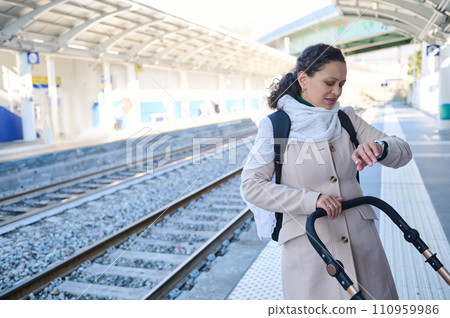 Young multi ethnic woman mother pushing baby stroller and checking time on wristwatch, feeling worried about train delay 110959986