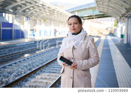 Young serious woman waiting train on platform of railway station. She is checking mobile application on her smartphone, booking online tickets and using the internet concept of mobile railway station Young serious woman waiting train on platform of railway station. She is checking mobile application on her smartphone, booking online tickets and using the internet concept of mobile railway station 110959989