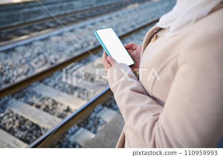 A smartphone with white blank touch screen with copy ad space, in the hands of a woman standing on the train platform A smartphone with white blank touch screen with copy ad space, in the hands of a woman standing on the train platform 110959993