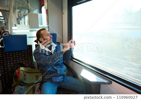 Cheerful young adult man in casual denim, gazing out the window during his morning commute on an urban light rail train 110960027