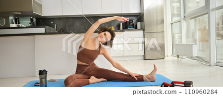 Workout and lifestyle concept. Portrait of fit, healthy woman stretching body at home on yoga mat, warm-up before training, doing exercises in bright room 110960284
