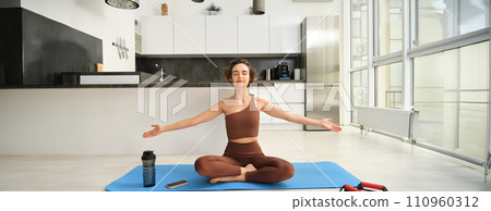 Mindfulness and meditation. Young relaxed fitness woman practice yoga at home, sitting in meditation pose, lifting hands up to the air and doing breathing exercises on rubber mat 110960312