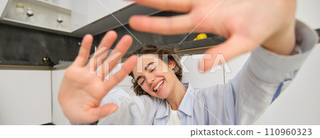 Lifestyle and people concept. Young happy woman hides from camera, extends hands forward, laughs and smiles carefree, spends time at home with joy Lifestyle and people concept. Young happy woman hides from camera, extends hands forward, laughs and smiles carefree, spends time at home with joy 110960323