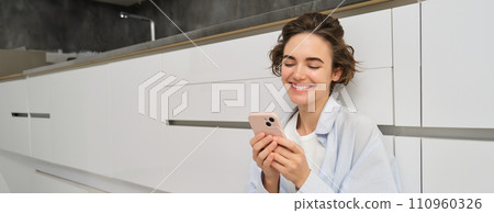 Portrait of cute smiling woman, sitting on floor at home with crossed legs, looks at mobile phone, chats on smartphone app, watches videos in application Portrait of cute smiling woman, sitting on floor at home with crossed legs, looks at mobile phone, chats on smartphone app, watches videos in application 110960326