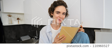 Portrait of brunette girl with short hair, holds planner, reads journal book on kitchen counter, enjoys leisure time at home Portrait of brunette girl with short hair, holds planner, reads journal book on kitchen counter, enjoys leisure time at home 110960327