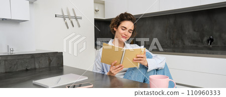 Smiling beautiful woman, sitting with notebook in kitchen, reading notes, studying, doing homework 110960333