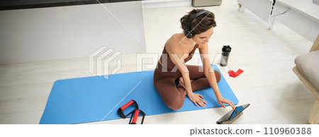 Fitness girl in rubber mat, looking for exercise tutorial, follows workout app instructions on digital tablet, listens music in headphones, uses rubber resistance band and plank equipment Fitness girl in rubber mat, looking for exercise tutorial, follows workout app instructions on digital tablet, listens music in headphones, uses rubber resistance band and plank equipment 110960388