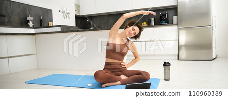 Young woman looking at digital tablet, connects to stretching fitness online class, workout, sits on yoga rubber mat at home, follows gym instructor exercises remotely 110960389