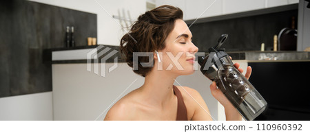 Portrait of fitness woman, young sportswoman at home, drinks water from bottle after workout, training exercises, takes a break after pilates, yoga training, stays hydrated 110960392