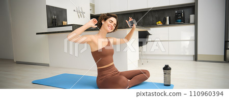 Active lifestyle and health. Happy young woman does workout, sits and dances with smartphone in hands, listens music in wireless earphones during fitness training session, yoga on floor mat 110960394