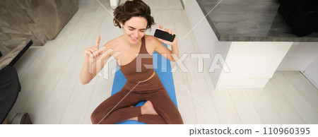 Top view of young active woman, healthy girl stretching, doing workout at home, holding smartphone and listening music in headphones while workout at home, does fitness, pilates yoga on floor mat 110960395