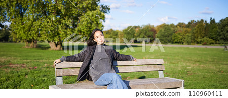 Portrait of young woman in outdoor clothes, sitting on bench relaxed, smiling and enjoying view on green park 110960411