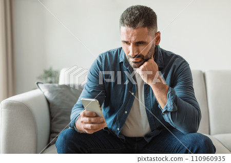 Thoughtful middle aged man doom scrolls through phone at home 110960935