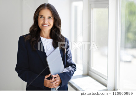 Mature woman entrepreneur holding laptop, posing by window 110960999