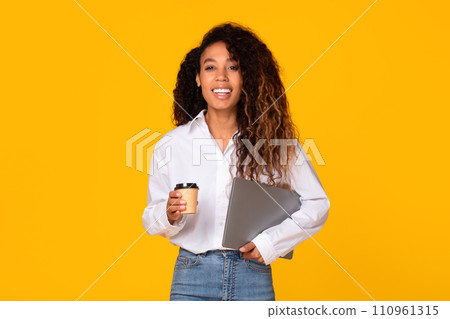 Happy black woman holding laptop and coffee on yellow background Happy black woman holding laptop and coffee on yellow background 110961315
