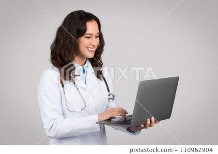 Happy caucasian lady doctor therapist in white coat typing on laptop, isolated on gray background studio shot 110962904