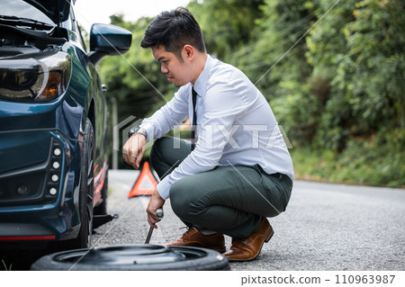 Experienced Asian engineer changing car wheel on the highway. Handsome businessman in white shirt watches the repair process. Skilled mechanic providing roadside assistance. Experienced Asian engineer changing car wheel on the highway. Handsome businessman in white shirt watches the repair process. Skilled mechanic providing roadside assistance. 110963987