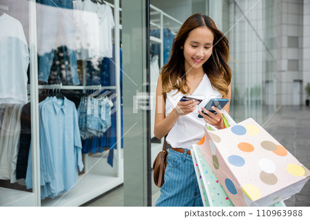 Young woman multitasking with shopping bags, smartphone, and credit card while browsing an online store. Convenience at her fingertips Young woman multitasking with shopping bags, smartphone, and credit card while browsing an online store. Convenience at her fingertips 110963988