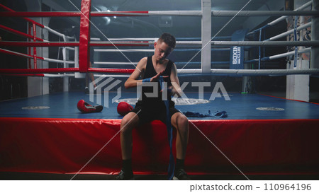 Young boxer sits near boxing ring and wraps his hands Young boxer sits near boxing ring and wraps his hands 110964196